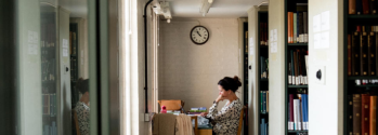 A University of Cambridge student reading a book next to bookcases under a clock at the Cambridge University Library. Credit: Alice the Camera/Cambridge University Library. Copyright: University of Cambridge (September 2019)